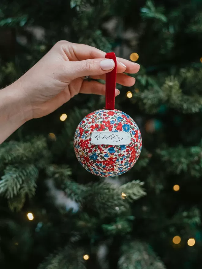 Personalised bauble with name held in front of a tree.