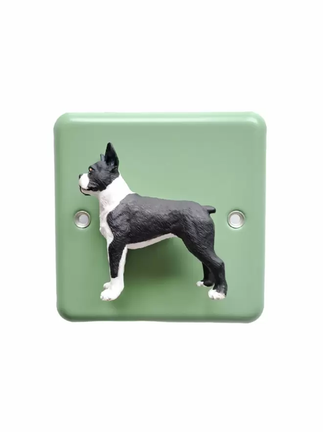 A boston terrier dog dimmer light switch. The light switch has a pastel green steel metal epoxy coated light switch plate, the boston terrier dog  is black and white and is made of plastic. The light switch is brand is Candy Queen Designs and Varilight. The boston terrier dog light switch light switch is on a plain white background.