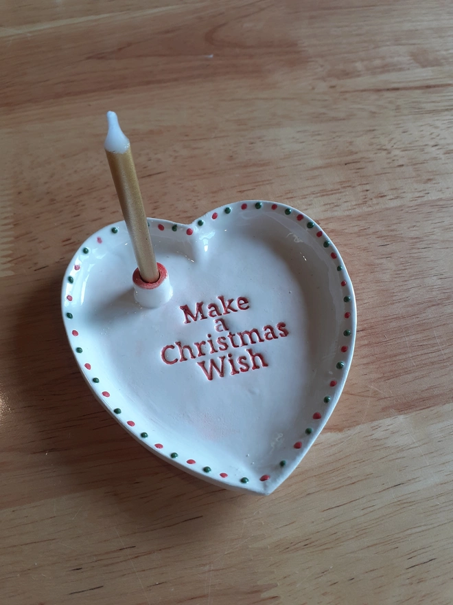 Handmade white ceramic heart-shaped Christmas Wish Dish with red lettering and alternating red and green dots, displayed beside a small box containing a gold candle and matches.