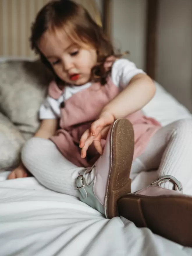 little girl sitting on a bed wearing Pip and Henry lilac T bar shoes