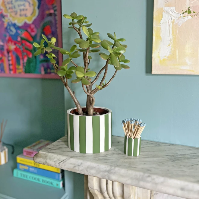 Green and white striped jesmonite pot with green plant inside. Positioned on a fireplace against a blue wall.
