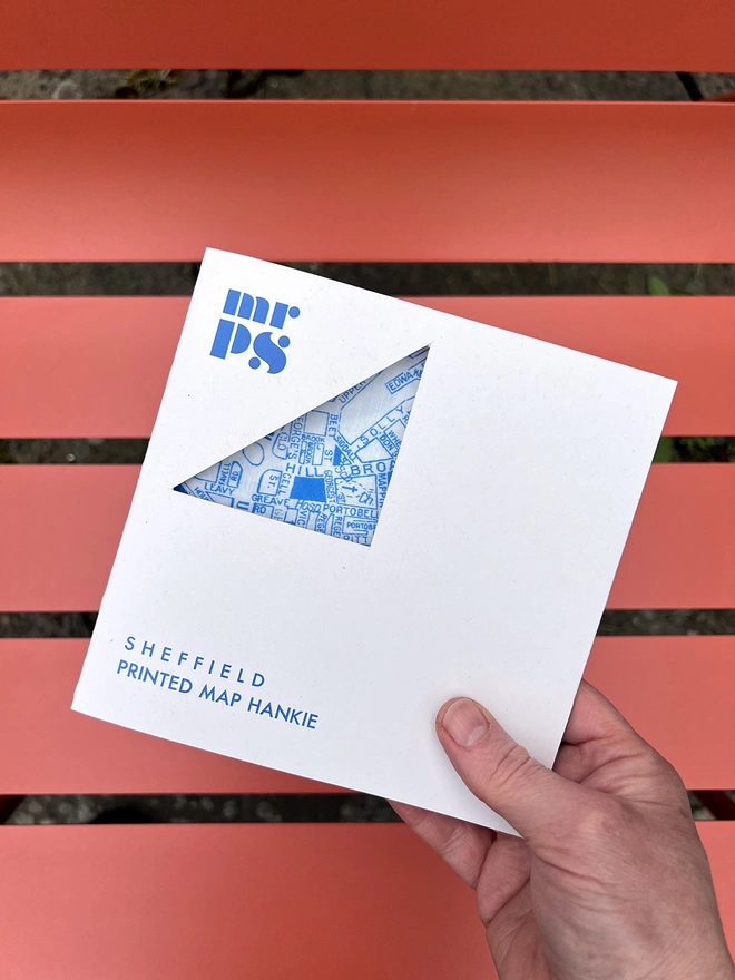 A white hand holds a Mr.PS Sheffield Handkerchief in a card packaging walletin front of an orange outdoor chair