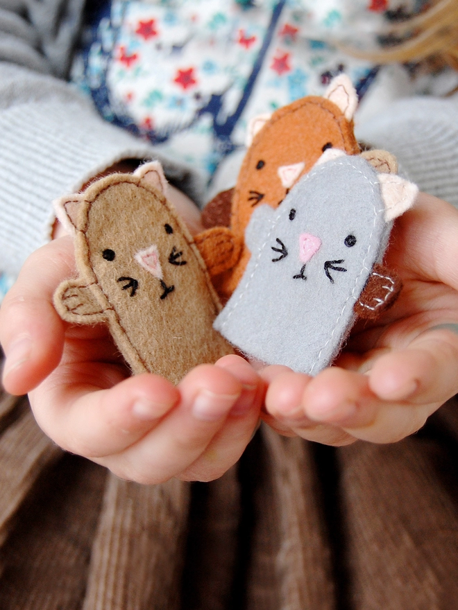A young child holds three homemade felt kitten finger puppets in her hands.
