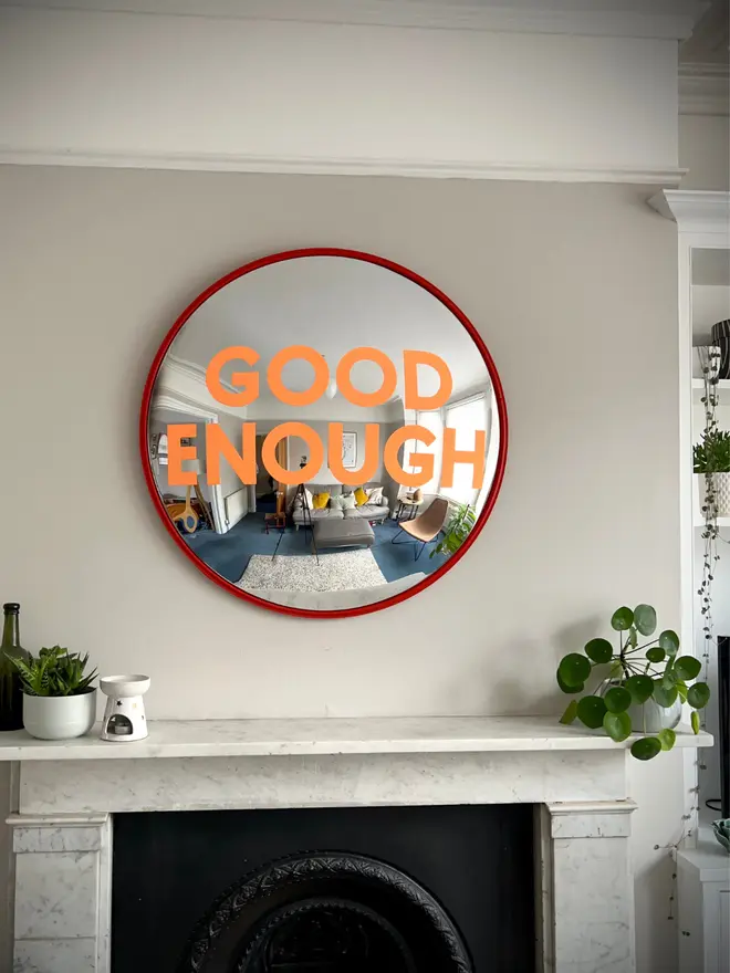 Round convex mirror with 'Good Enough' hand painted in orange in a red round frame, above a cream fireplace surrounded by house plants