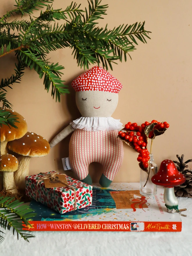Soft handmade mushroom-themed doll with a red spotted cap, embroidered smiling face, ruffled white collar, red-striped body, and small green feet.