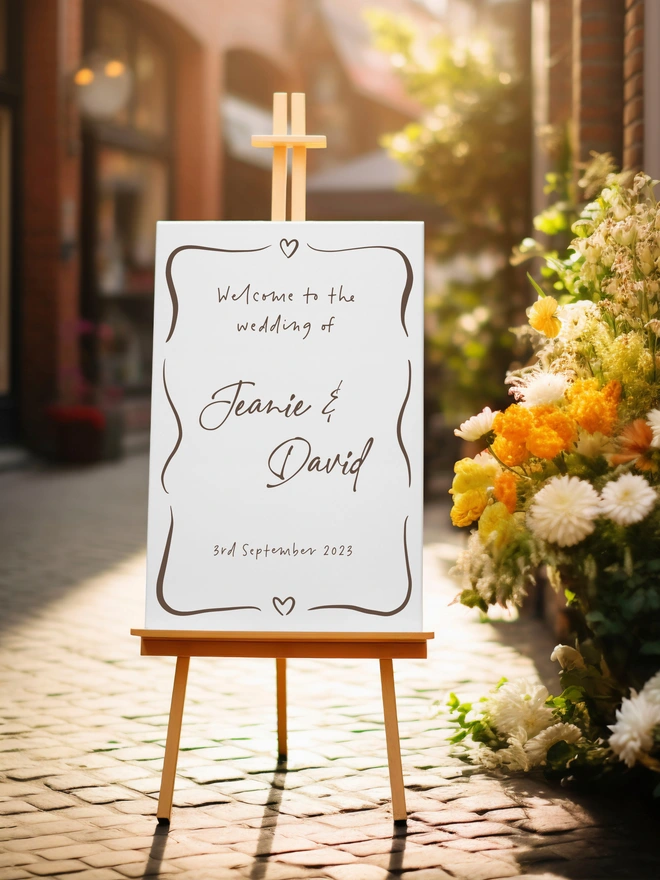 A white board on a wooden stand with the writing in brown of Welcome to the wedding of Jeanie and David on the 3rd September 2026 and at the top and bottom are hearts with some squiggles encasing the words. The stand is on a cobbled path with a an arrangement of yellow, white flowers and green foliage.