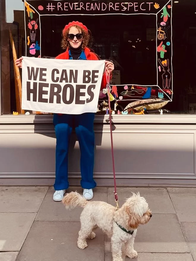Person with dog stood in front of a shop window that says #revereandrespect holding a white tea towel with black text that says We Can Be Heroes