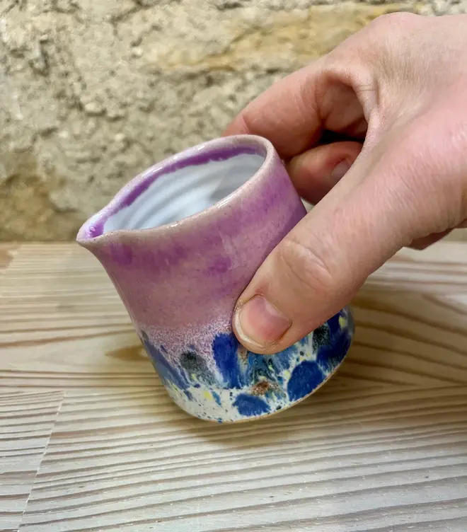 Shoreline Tiny Jug, a colourful tiny jug placed on a wooden surface. It sits in front of a brick wall backdrop. In the image its being poured. 