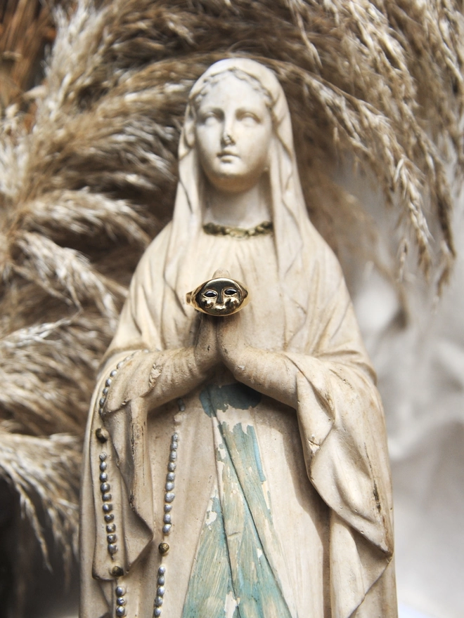 Image of am antique Virgin Mary statue with an 18 carat gold moon face ring with sapphire eyes balancing on her praying hands, in the background there us dried pampas grass