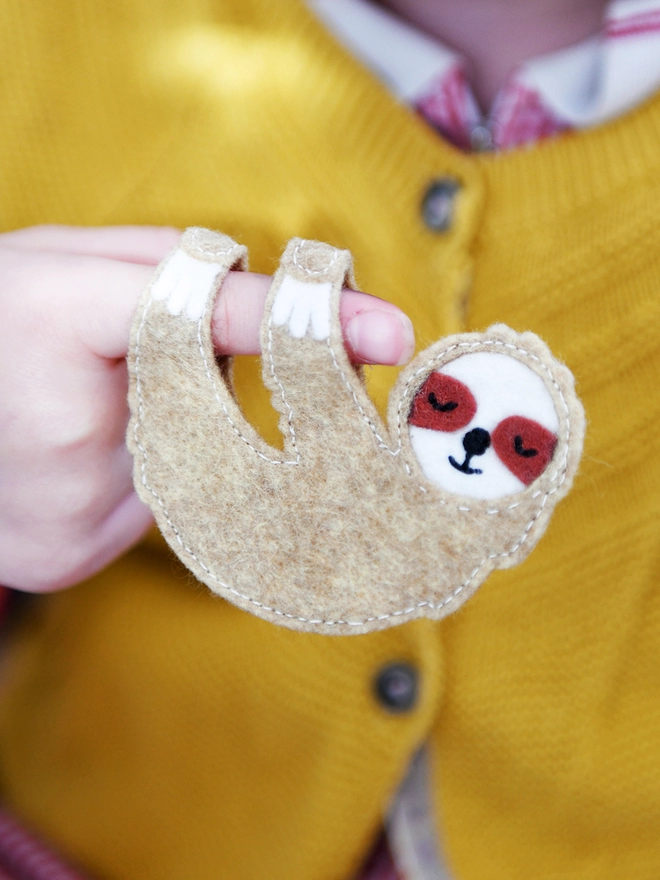 A handmade sloth finger puppet made from beige and white felt is hanging from a young child's finger.