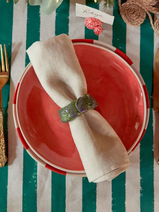Handmade ceramic Brussels sprout napkin ring holding a natural linen napkin on a coral-red plate, styled on a green-and-white striped Christmas table with gold cutlery and festive foliage.