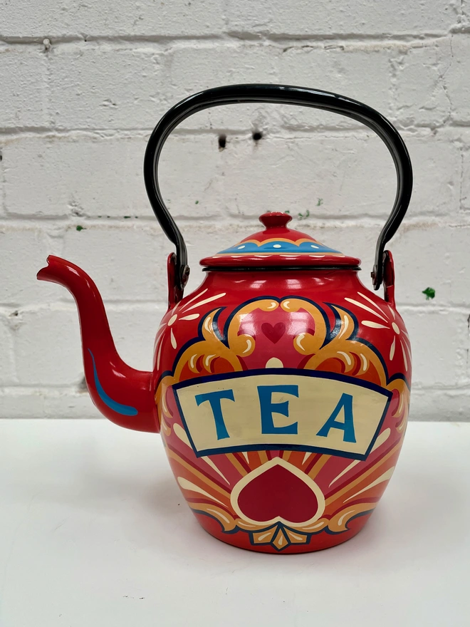 Red vintage enamel teapot with folding pouring handle, hand-painted with a traditional fairground inspired design in pink, orange and yellow. 'Tea' is painted in a banner across the centre.