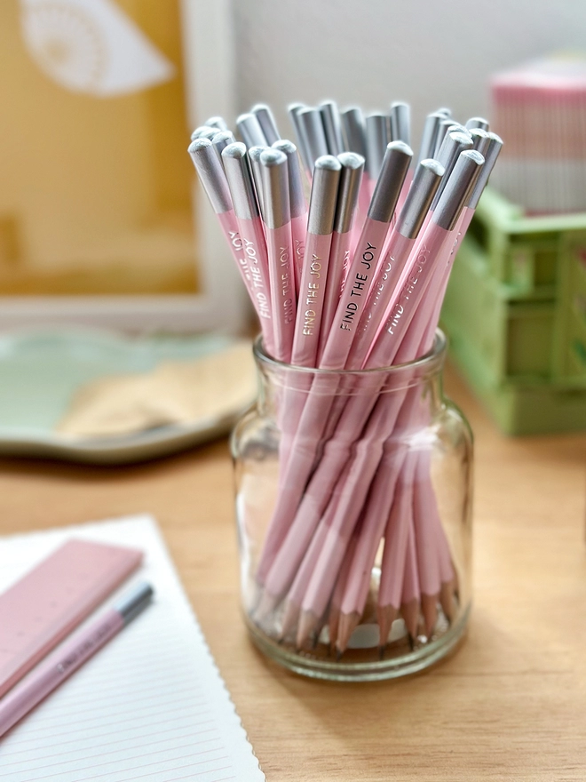 A glass jar full of pink pencils stands on a wooden desk with various stationery items around it.