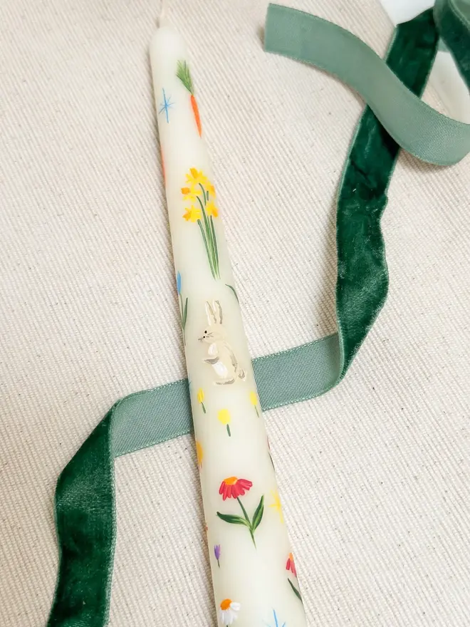Spring Fun Candle, a white spring candle lying on a plain surface accompanied by a green ribbon. It's floral design can be seen wrapping around it. 