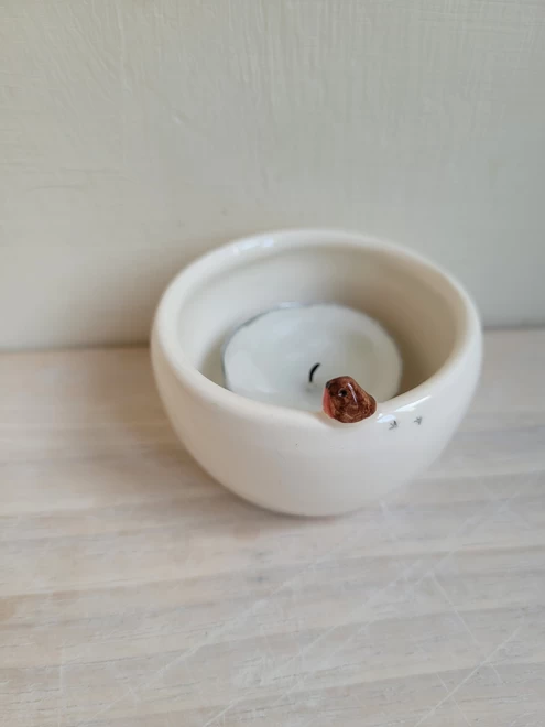 a small ceramic tealight candle holder with a small modelled robin attached to the rim with tiny bird prints . There is a white candle inside it is on a light wooden surface 