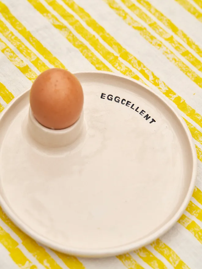 Egg And Soldier Plates, a plate with an integrated egg cup holding an egg placed on a yellow striped surface. The wording reads 'eggcellent' 