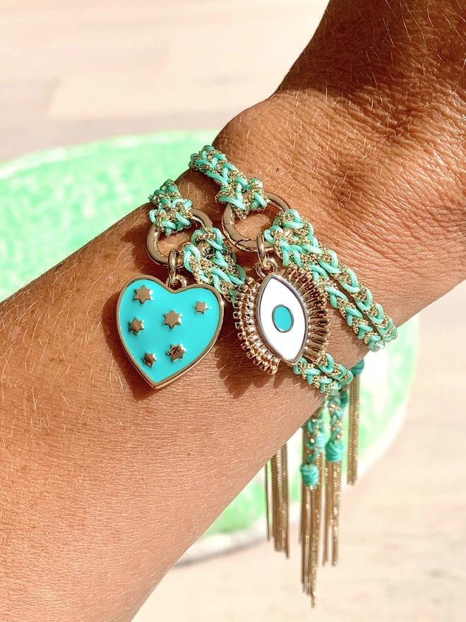 Woman’s wrist wearing a gold chain and turquoise silk friendship bracelet threaded with a turquoise heart charm
