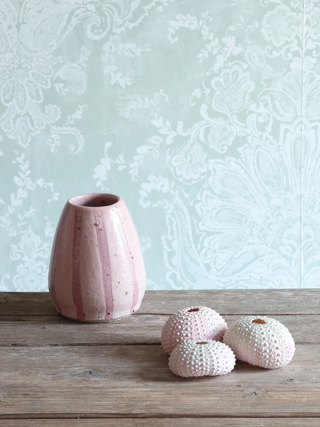 Handmade Ceramic Stoneware Small Vase in Pink sat on a wooden surface alongside a collection of three pink sea urchins. 