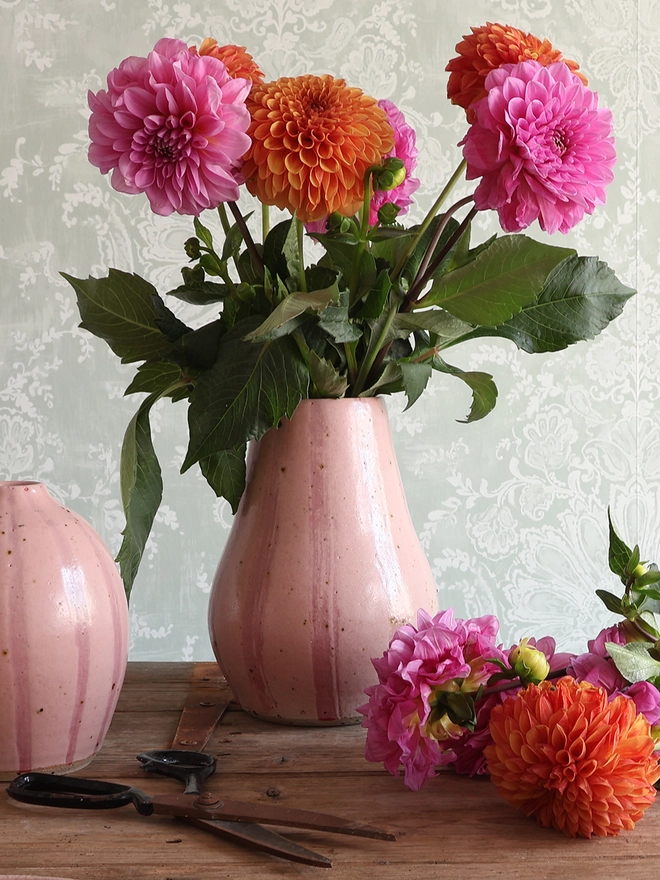 A Handmade Ceramic Stoneware Large Vase in Pink Stripe filled with a bouquet of pink and orange flowers. A medium, rounded vase in the same style sits next to the larger vase, along with additional flower stems and a pair of scissors. 