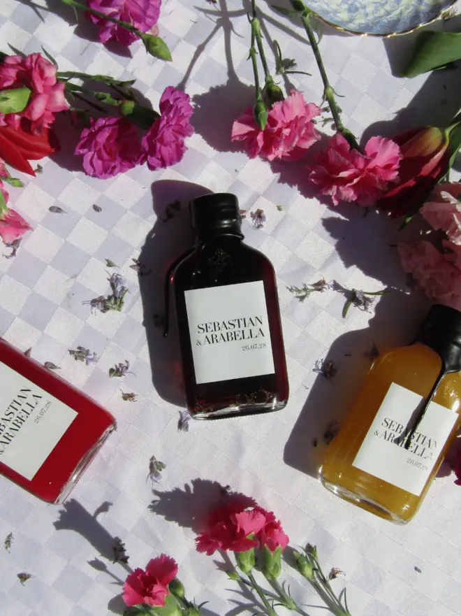 multiple glass bottles of various coloured cocktails with white label with names of wedding couple, against floral backdrop
