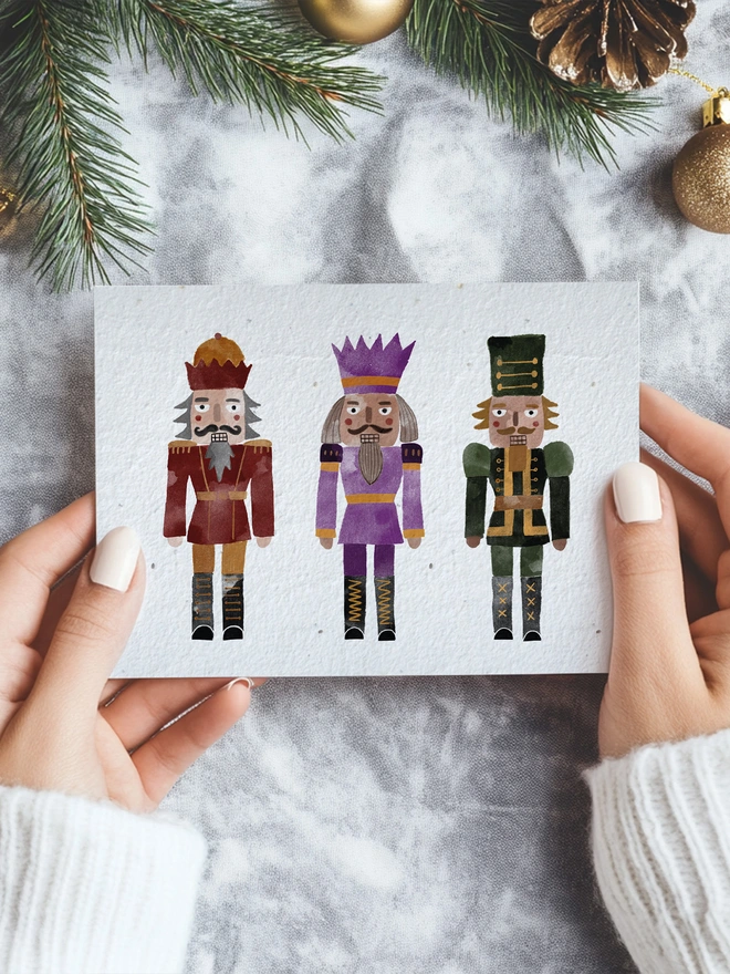 A pair of hands holding a Christmas card with three illustrated watercolour nutcracker in a Christmas scene
