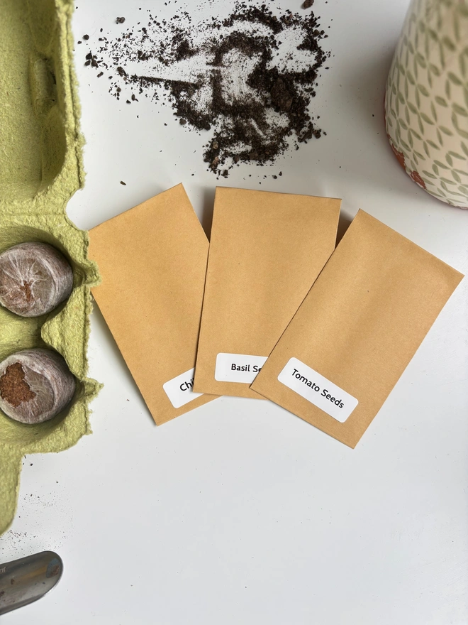 three brown kraft seed packets labelled on a white background. Messy soil above them with an open egg box with compost discs next to it.