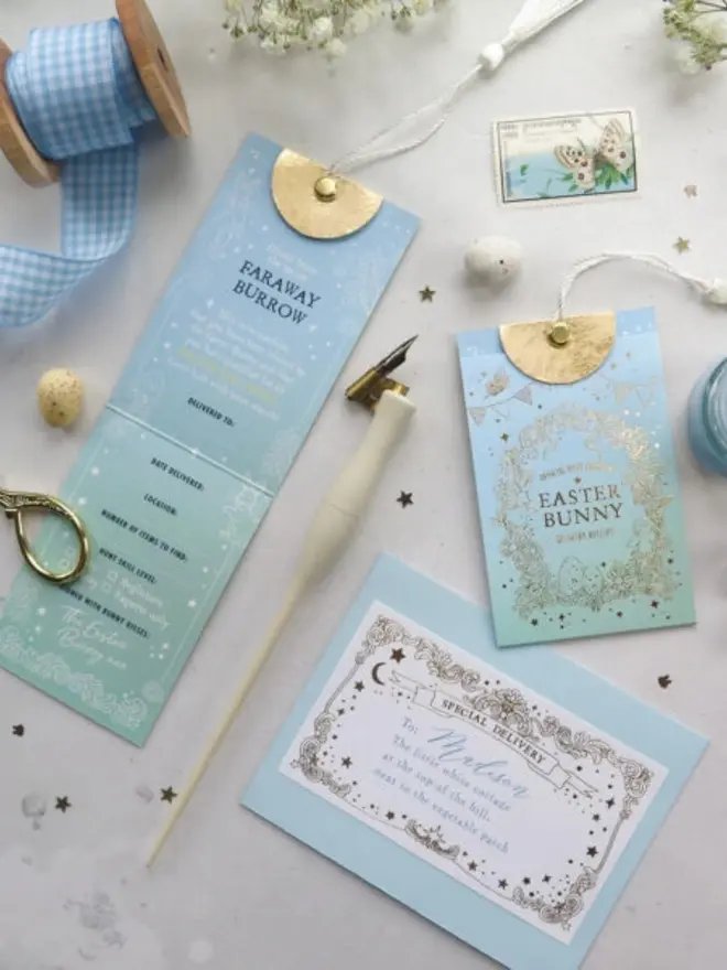 The fold-out egg hunt card, gold foil pouch and a blue envelope addressed to Madison laid out together. A dip pen, blue gingham ribbon and gold scissors complete the scene.