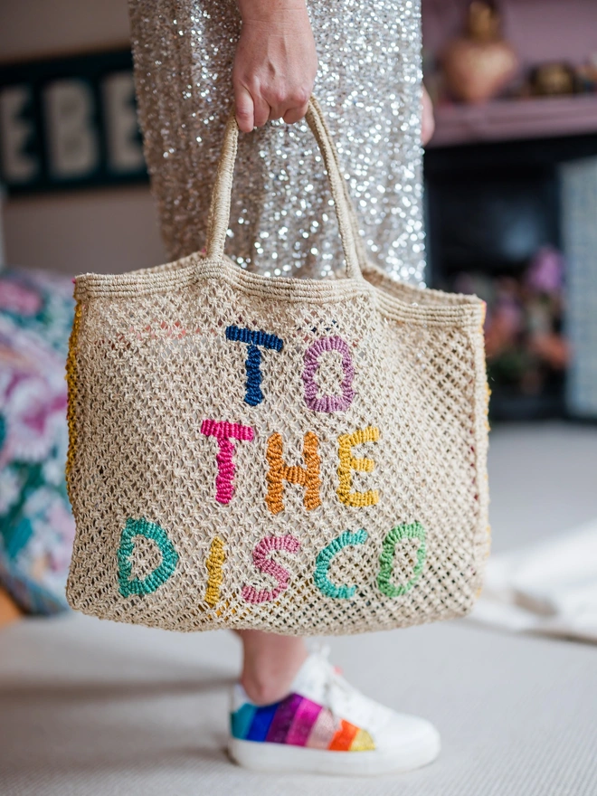 A person holding a "To the Disco" jute bag with colourful trainers in an indoor setting