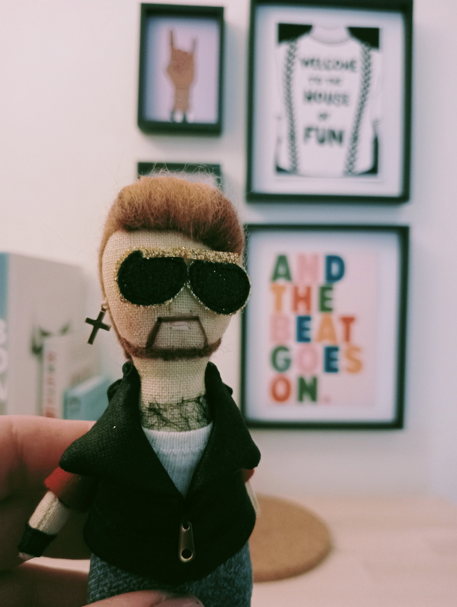 George Michael mini decorative doll wearing faith era leather jacket and tight jeans with black painted boots crucifix earring and metallic aviator glasses held in a left hand in front of a gallery of music themed prints