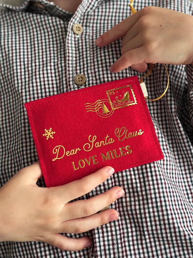 A child wearing a gingham shirt is holding a handmade red felt Christmas envelope that reads "Dear Santa Claus, Love Miles" in gold writing with a golden sleigh stamp in the top right hand corner.