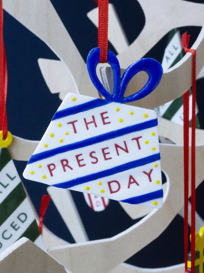 Ceramic porcelain Christmas decoration with The Present Day written on the side. A white present-shaped Christmas decoration with red lettering, decorated with blue stripes and yellow spots, and hanging from a plywood Christmas tree.