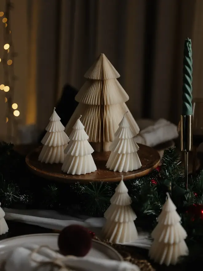 White Christmas Tree Warm Candles (Set Of 2). Cream Christmas tree candles with pleated, layered tiers grouped on a wooden board around a paper honeycomb tree, with pine garland and a twisted green taper candle. The minimalist sculptural candles make an elegant, understated festive centrepiece.