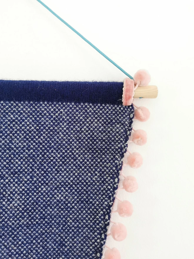 A product image showing the detail of the reverse of a pennant flag wall hanging. The back of the banner is knitted into a navy and white birdseye check. Here you can also see the wooden doweling, blush pink velvet pom pom trim, and bright teal nylon cord.