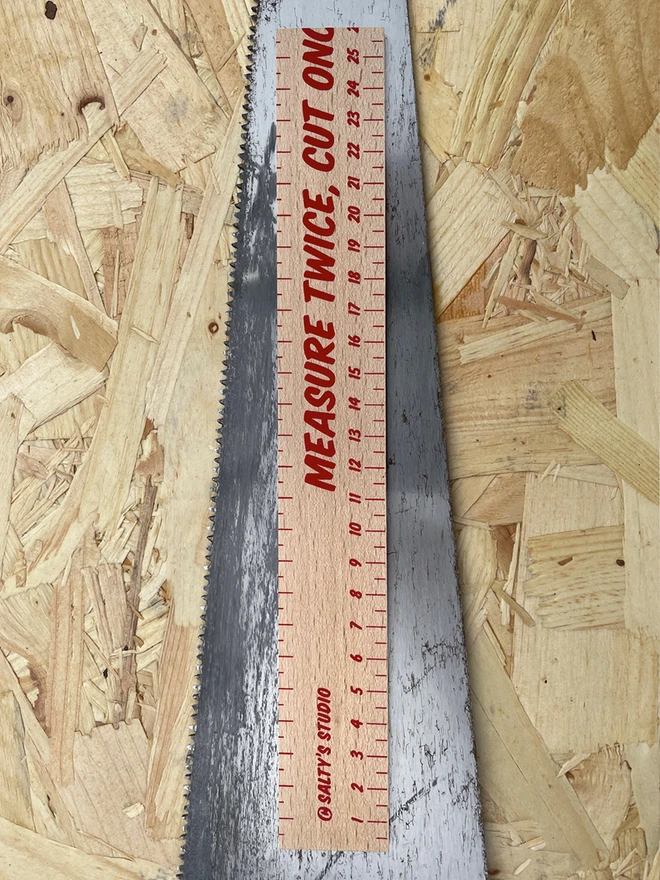 Measure Twice Cut Oncew ruler with part of the phrase cut off, for humour! 26cm screenprinted on once side. Sat on a saw, all on a chipboard background.