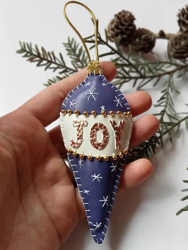 A Hetty and Dave purple hand stitched leatherette retro inspired bauble with gold metallic lettering spelling out JOY on a white band, adorned with tiny gold beads
