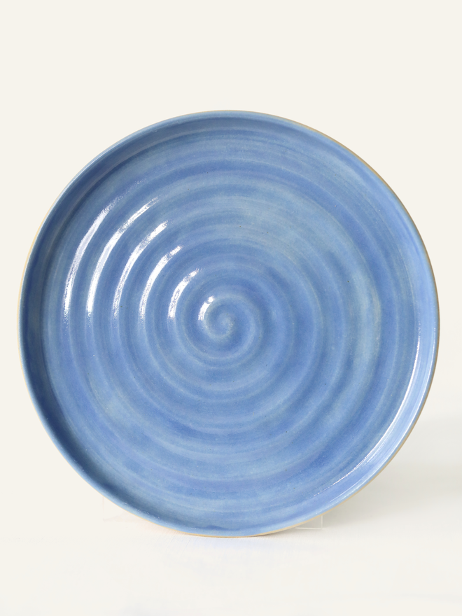 A handmade ceramic lunch plate in glossy pacific blue glaze, photographed from above against a plain light grey background, showing a natural concentric swirl texture from the wheel-throwing process