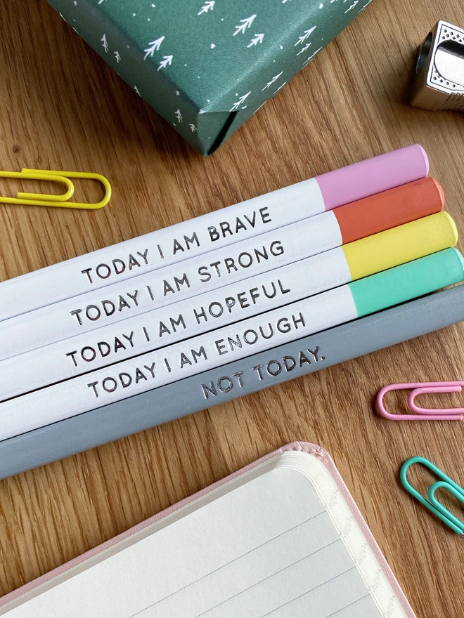 Five white pencils with pastel coloured tips and positive messages stamped in silver foil on the sides, lay on a wooden desk.