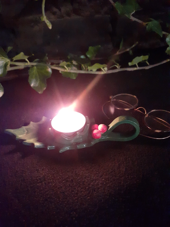 Handmade ceramic holly leaf tealight holder glowing with a lit candle, styled against a dark background with natural ivy.