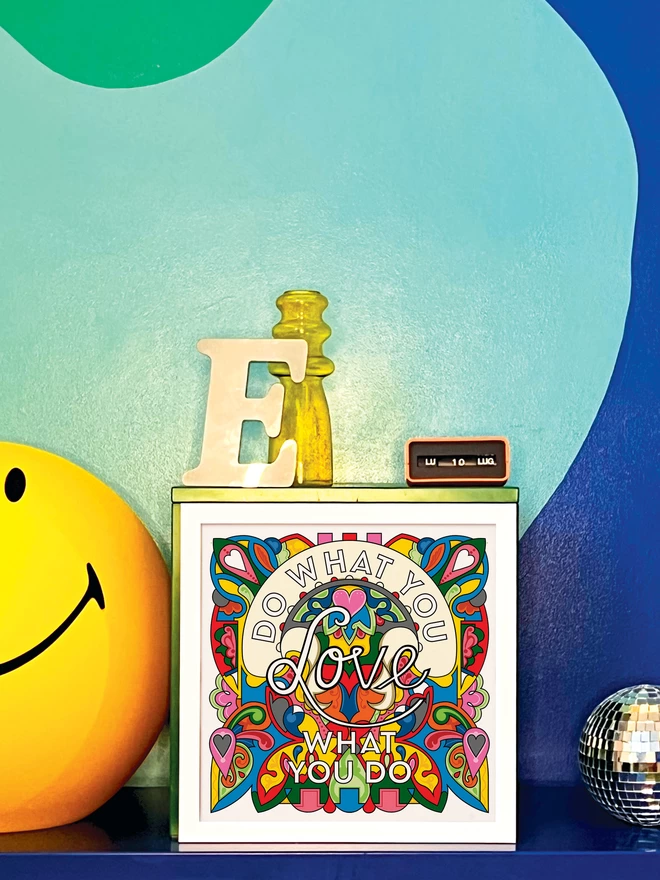 “Do what you love, love what you do” is written over a vibrant, rainbow coloured illustration in a white frame propped against a turquoise and dark blue wall. Next to the frame is a disco ball,a letter ‘E’ ornament, a yellow glass vase, an orange Italian plastic calendar showing the date as ‘LU 10 LUG’ and a large light up yellow Smiley lamp.