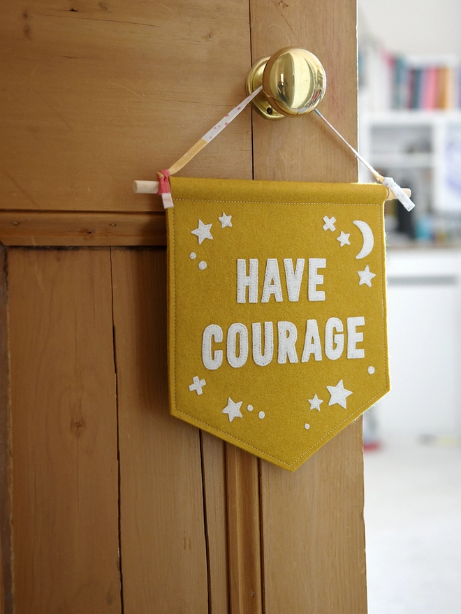 A mustard yellow banner flag, with the words Have Courage in white, hangs on a wooden door handle by it’s ribbon attached to a wooden dowel at the top of the flag.