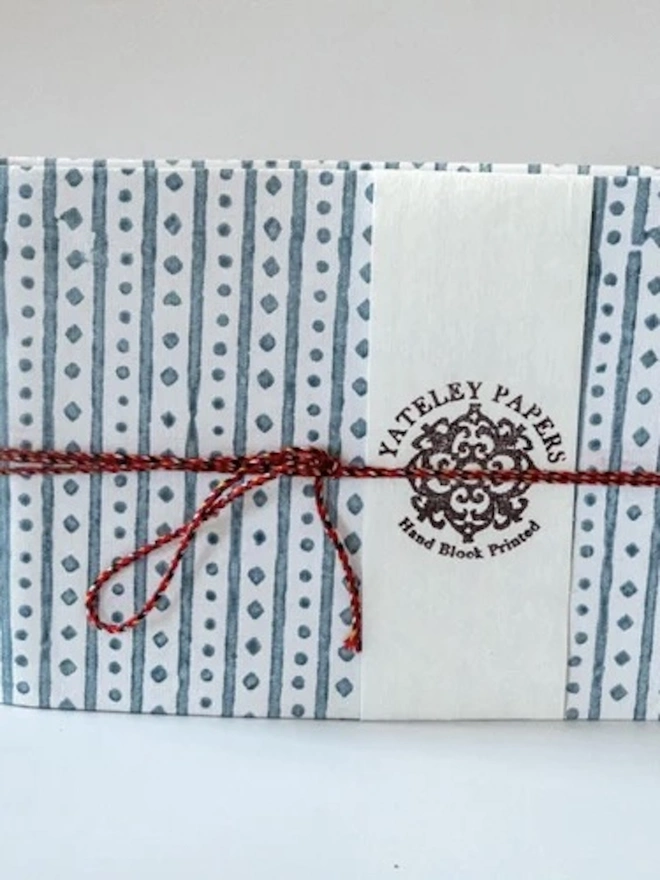 A5 Small Landscape Travel Watercolour Book. A Yateley Papers notebook covered in slate blue stripe and diamond-dot block-printed paper, finished with a cream branded band. A red braided cord bow adds a handmade touch to this artisan stationery gift.