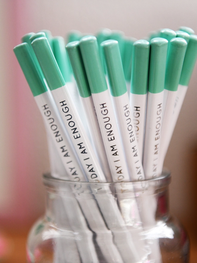 A glass jar full of white pencils with green tips and the words "Today I Am Enough" along the side of each one.