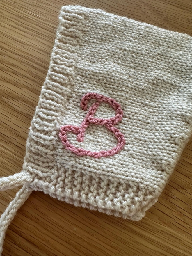 Pixie Bonnet. A cream pixie bonnet with a pink, embroidered ‘B’ on one side.