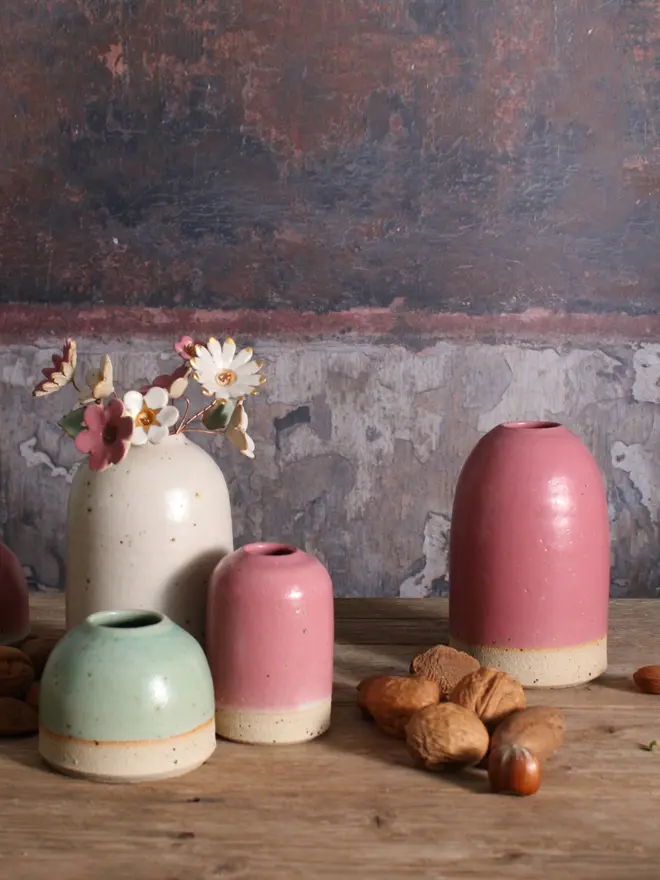 A cluster of our mini, small and medium ceramic vases in mint green, fuchsia and off-white. The medium vase holds a fuchsia and white ceramic flower bouquet with gold lustre detailing. The vases sit on a wooden surface alongside seasonal foliage and a dark textured backdrop