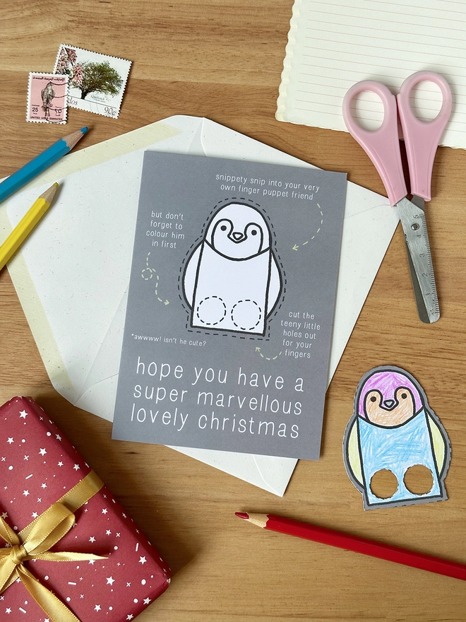 A colour in Christmas card with a penguin finger puppet design lays on a white envelope on a wooden desk.
