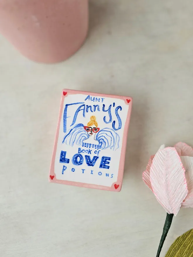  A charming ceramic trinket box designed to look like a vintage book, painted in shades of pink and red. The cover features the title "Aunt Fanny’s Book of Love Potions" in a playful, illustrated style. With its book-like shape and whimsical design, this decorative piece adds a touch of romance and mystery while offering a secret spot to store small treasures.