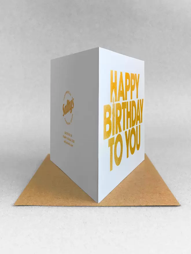 Happy Birthday to You landscape greetings card. The I of Birthday is a candle using negative space. Printed in capital letters, simple font printed in a gold ink on a white card. rear view showing the logo on the back and stood on a Kraft brown envelope made by Salty's Studio.