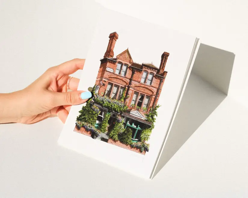 Nunhead Skehans Giclée Print (Unframed), a print of a pub being held by a woman above a plain surface. 