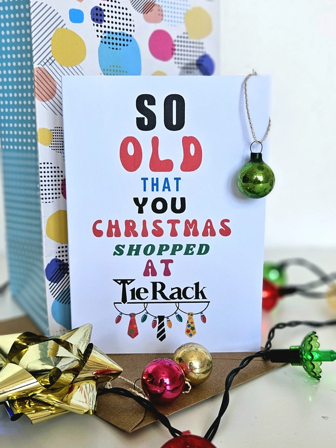 A photograph of the front of a humorous A6 greeting card illustrated using brightly coloured text and the retro shop logo. The text says so old you Christmas shopped at Tie Rack with a string of fairy lights and 3 colourful ties dangling from it
