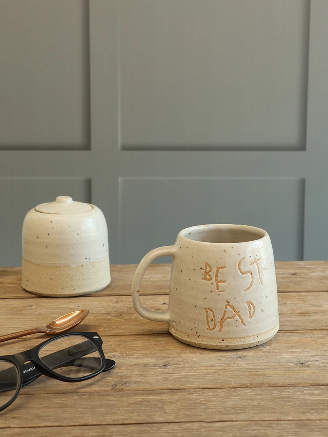 Personalised Handwriting Stoneware mug with the text 'Best Dad' in a child's handwriting on the front. The mug is sat on a wooden surface with a matching sugar pot, rose gold spoon and a pair of glasses. 